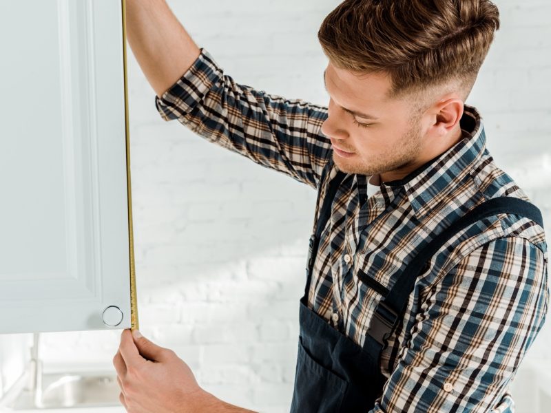handsome installer in overalls measuring white kitchen cabinet door