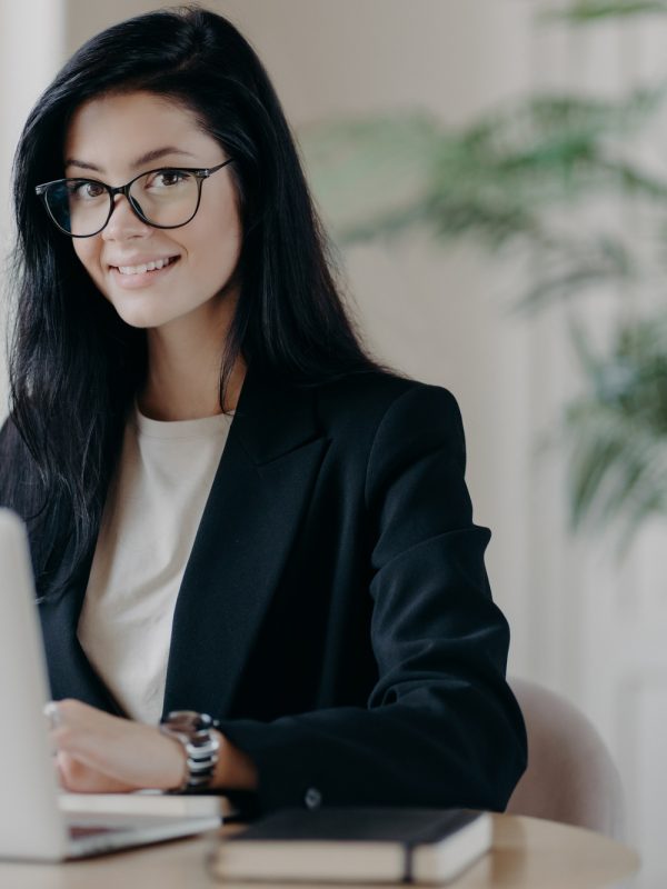 Smiling well dressed young woman secretary with dark hair, works on laptop computer at desktop